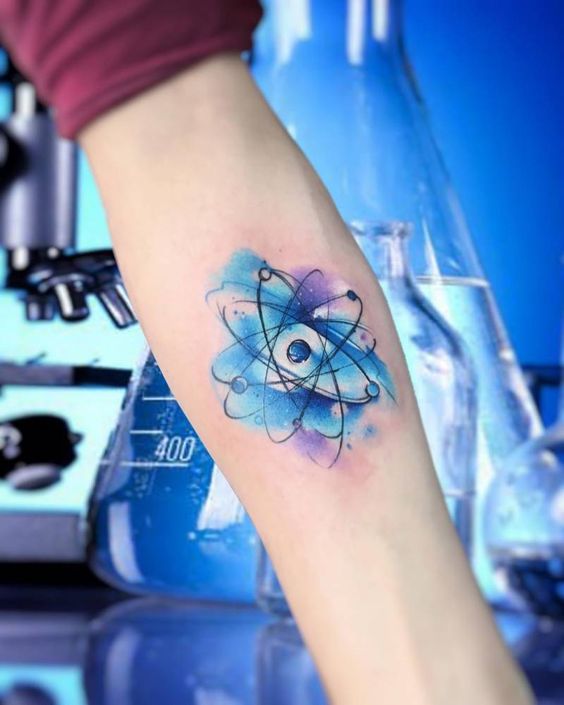 Tatuaje de un átomo de estilo acuarela, situado en el interior del antebrazo.